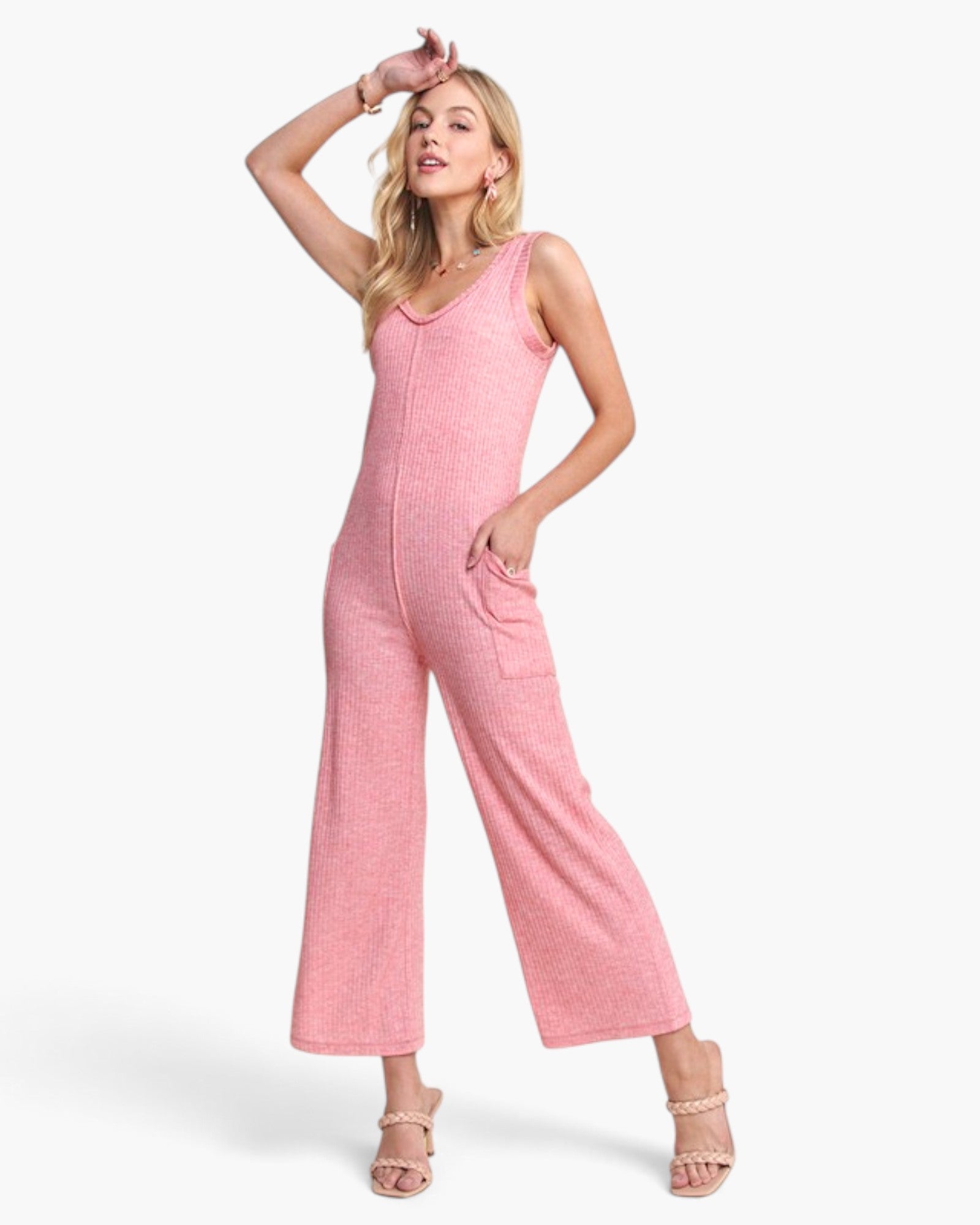 A woman modeling a pink sleeveless jumpsuit with wide legs and side pockets, paired with beige strappy heels, against a plain white studio background.