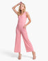 A woman modeling a pink sleeveless jumpsuit with wide legs and side pockets, paired with beige strappy heels, against a plain white studio background.