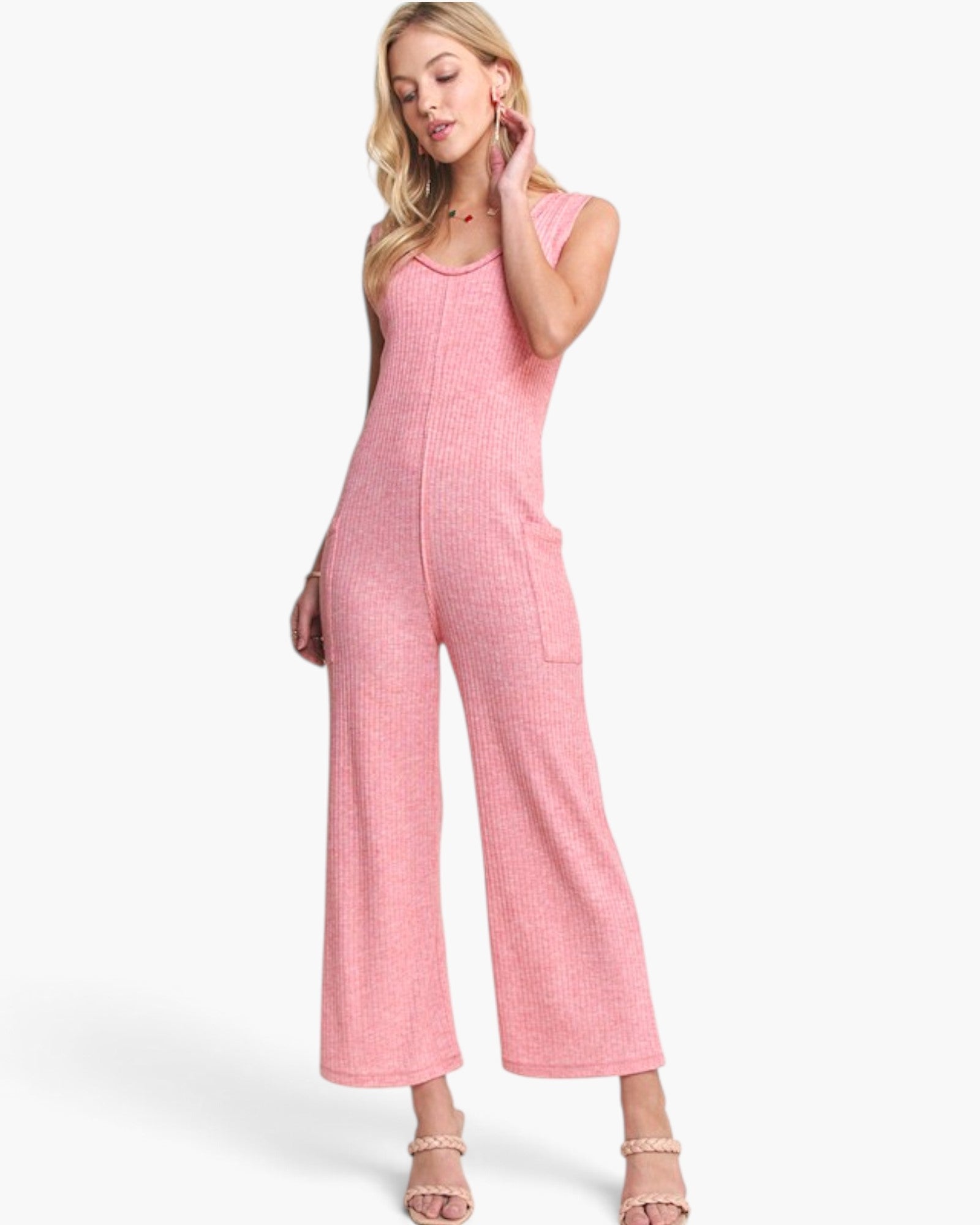A woman modeling a pink ribbed knit sleeveless jumpsuit with wide legs and front pockets, paired with beige strappy heels against a plain white studio background.
