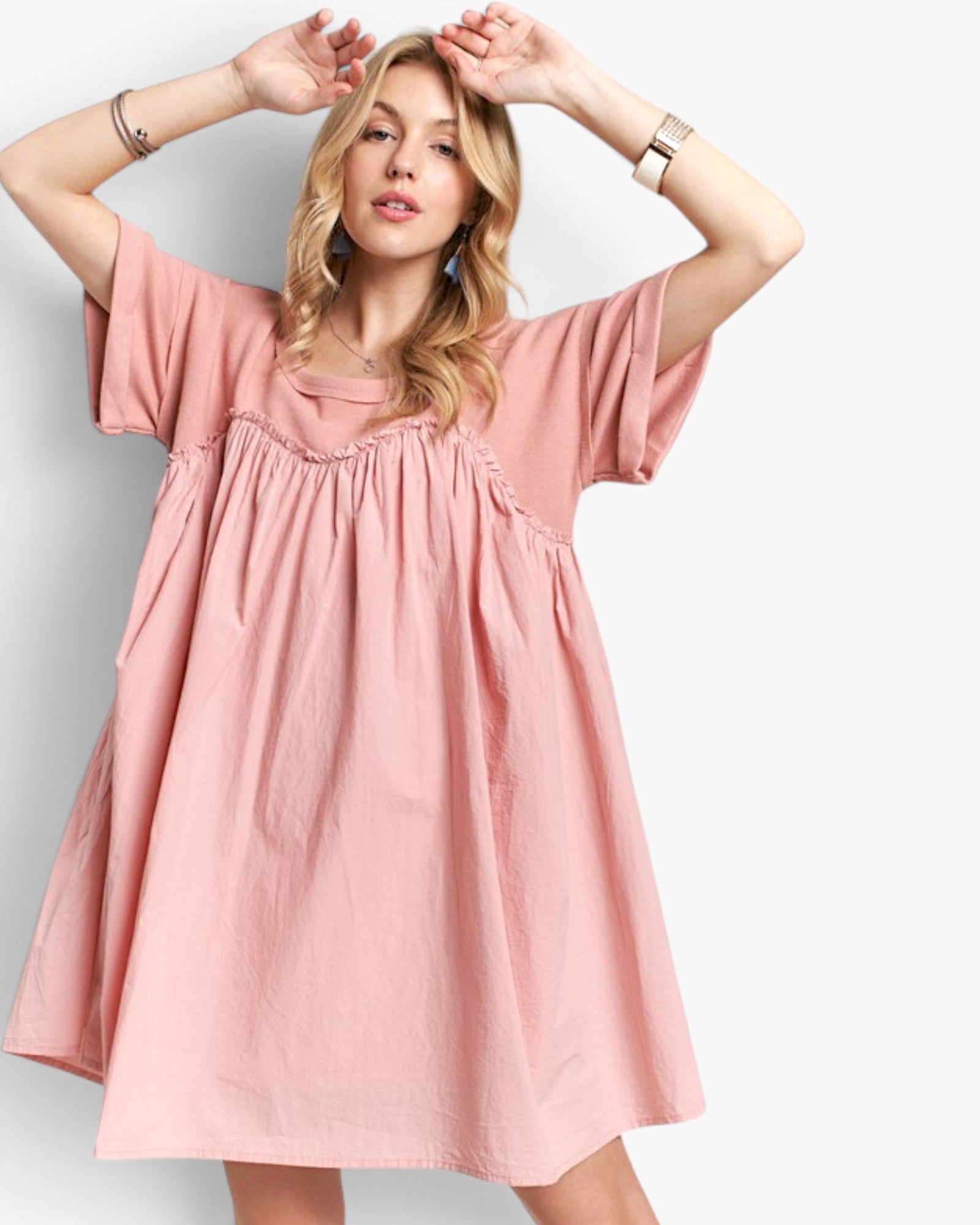 A woman modeling a dusty pink short-sleeve dress with a gathered smocked bodice and a loose, flowing skirt, set against a plain light gray background.