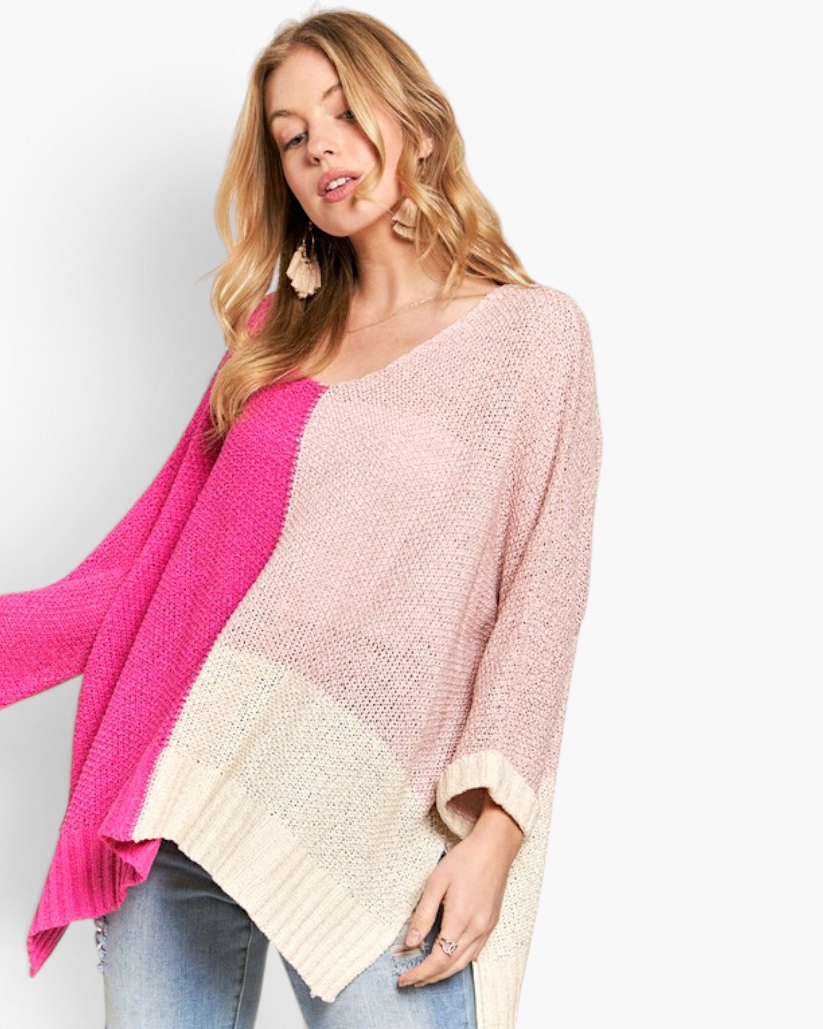 A woman wearing a chunky knit color-block poncho sweater in pink, cream, and bright magenta, paired with light blue jeans against a plain white background.