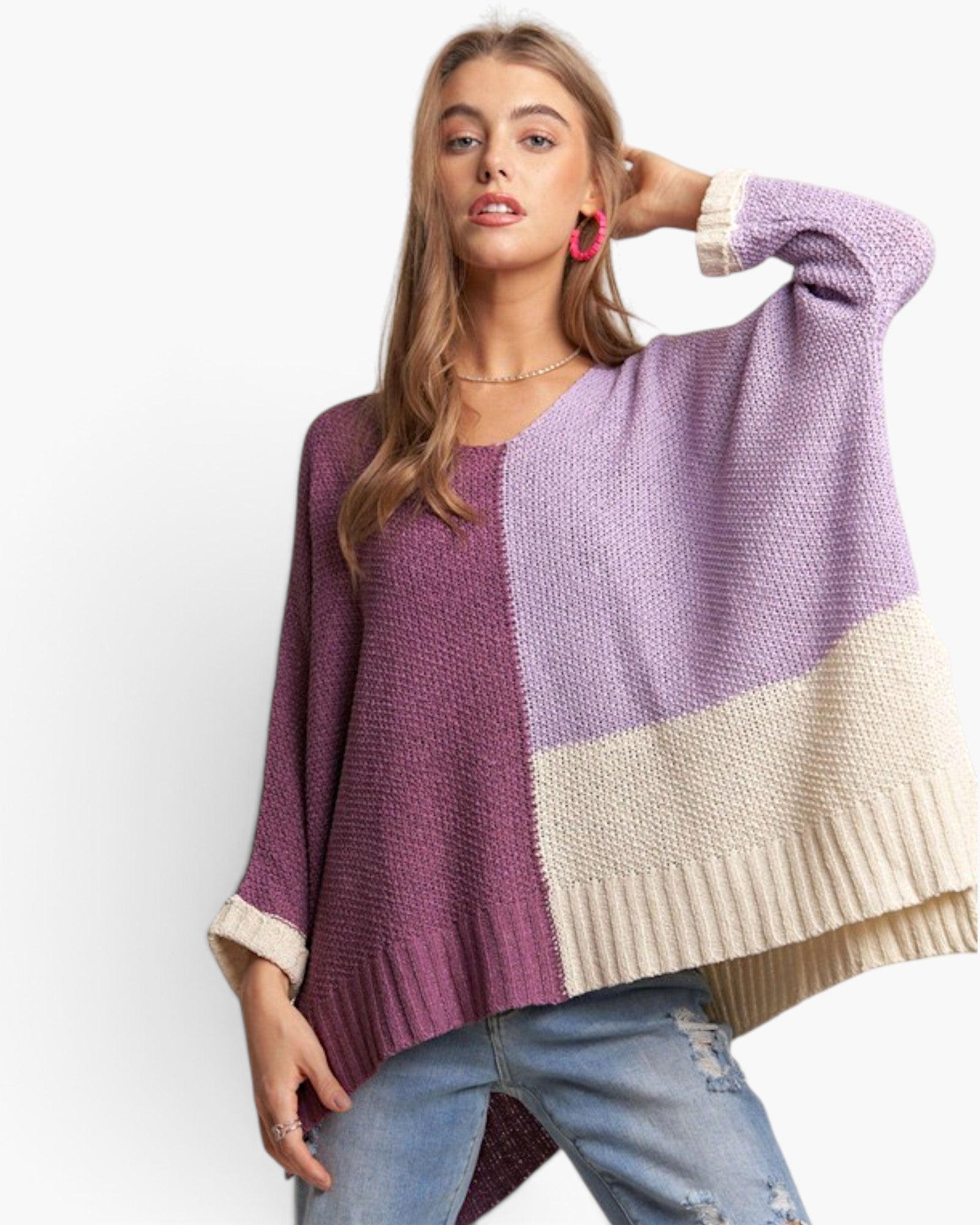 A woman modeling a chunky knit colorblock poncho sweater with a purple and cream color scheme, paired with blue jeans against a plain white background.