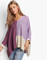 A woman modeling a loose, open-front knit poncho sweater with color-block panels in lavender purple and dusty mauve, plus cream ribbed trim and lower panels, paired with light blue jeans against a plain light gray studio background.