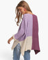 A woman wearing a chunky knit color-block poncho sweater with lavender, deep purple, and cream panels, paired with blue jeans, shown from a side-back angle against a plain white background.
