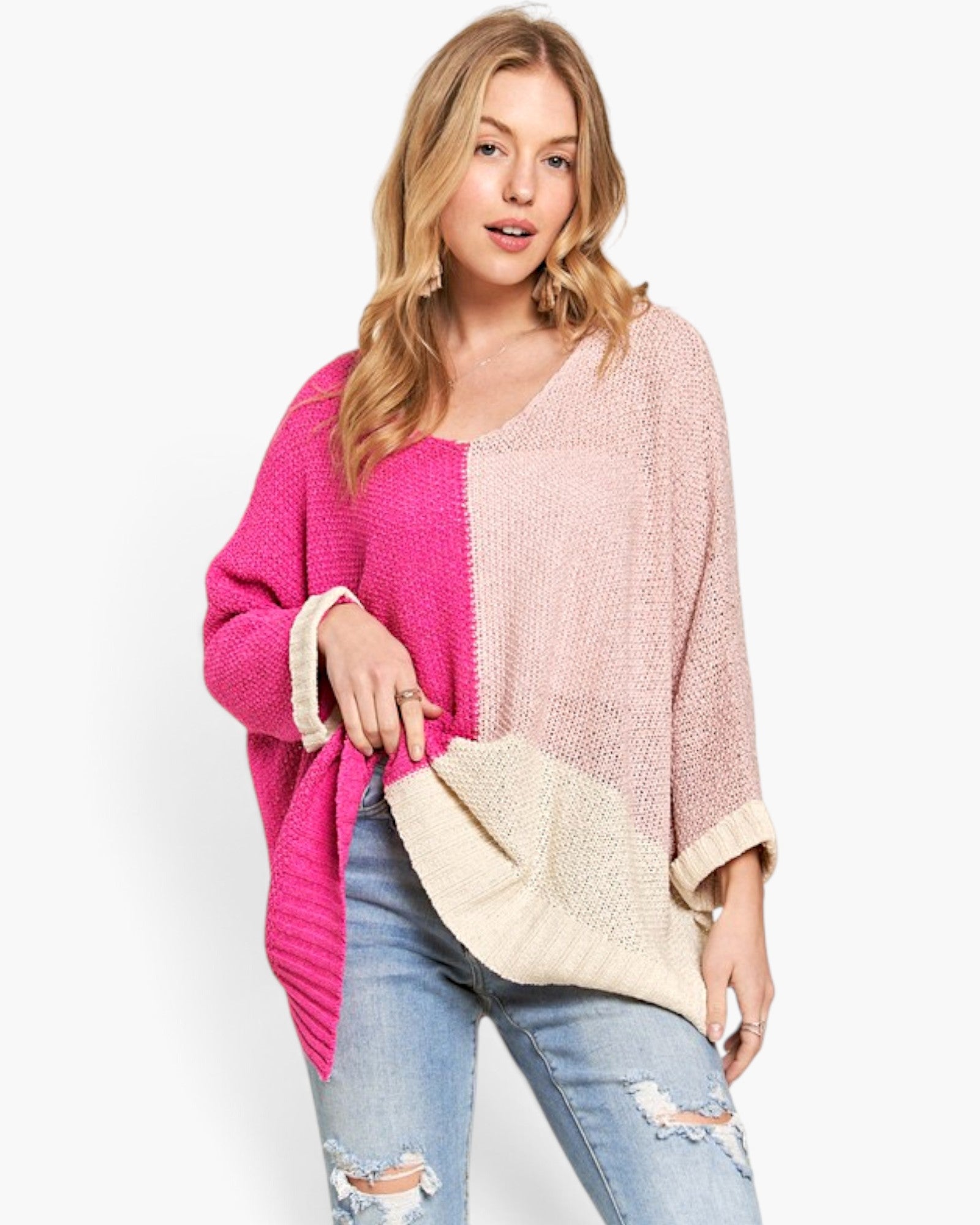 A woman modeling a color-block knit poncho sweater with bright pink, light pink, and cream panels, paired with distressed blue jeans against a plain white background.