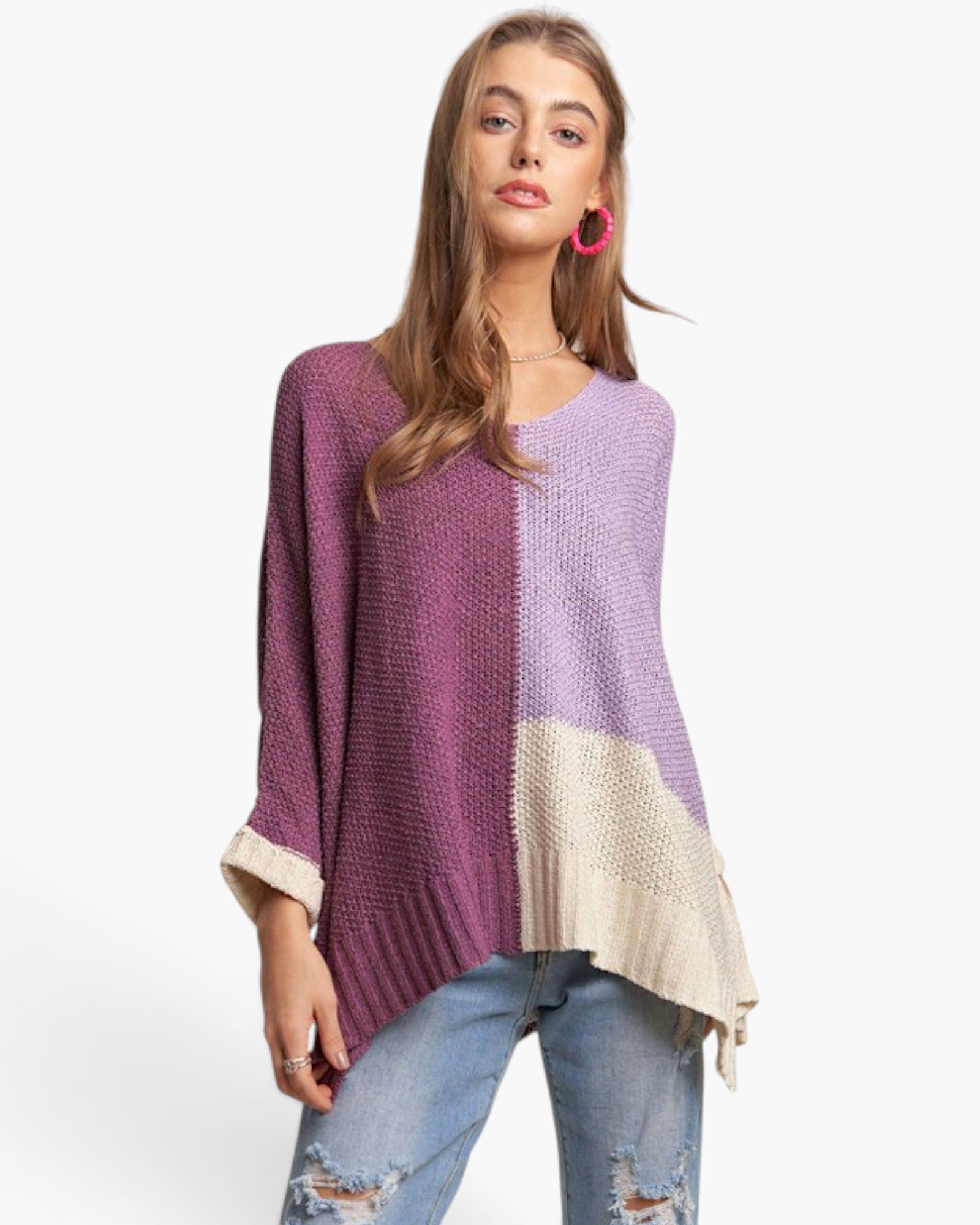 A woman modeling a color-block knit sweater with a split purple and lavender design, a cream-colored lower panel, and rolled cuffs, paired with distressed blue jeans against a plain white background.
