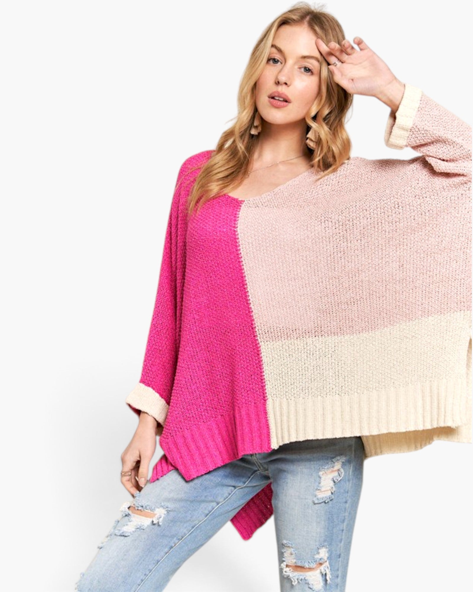 A woman modeling a chunky knit colorblock sweater with bright pink, dusty pink, and cream panels, paired with distressed blue jeans against a plain white background.