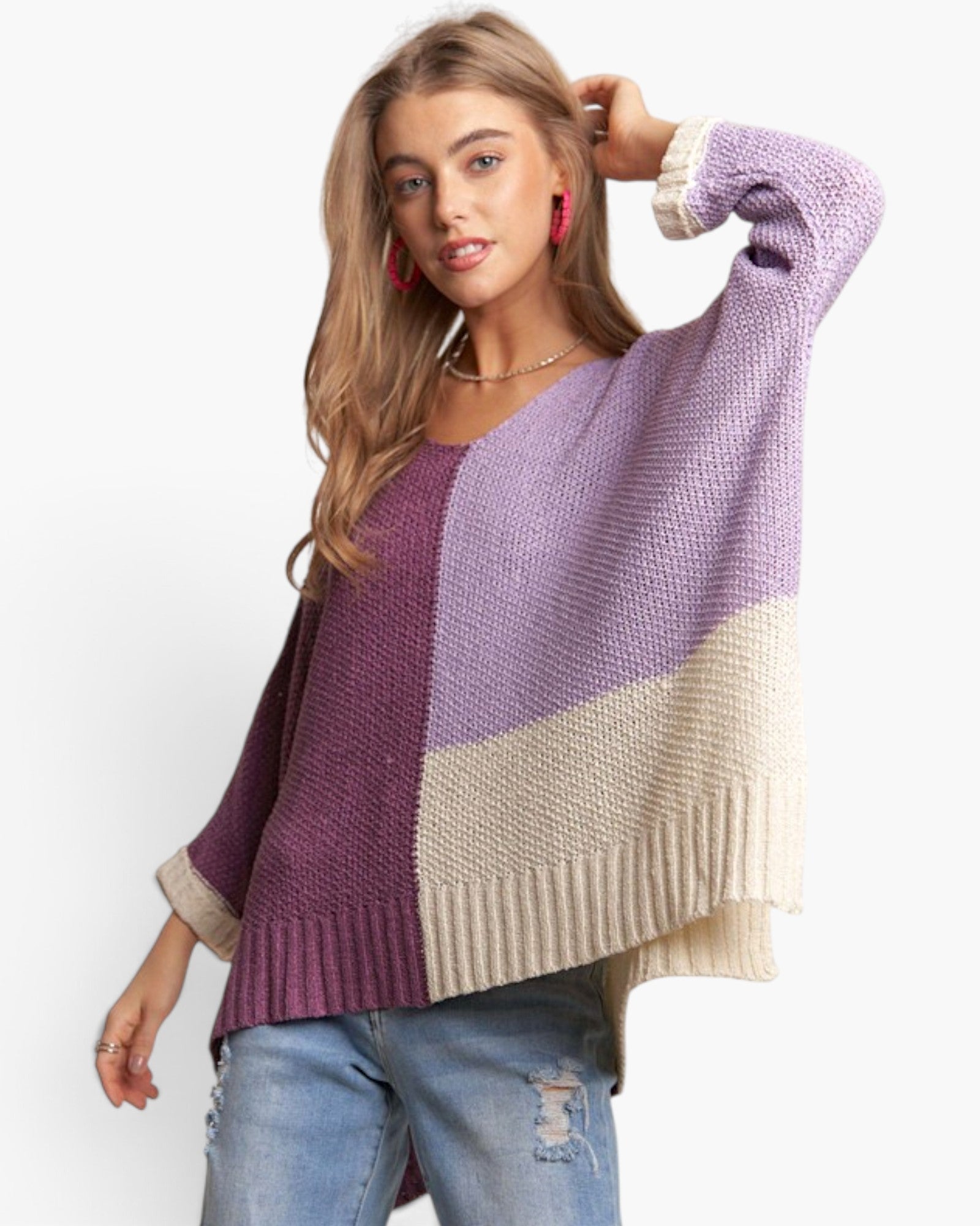 A woman modeling a chunky knit color-block sweater with lavender purple, deep plum, and cream panels, paired with distressed blue jeans against a plain white background.