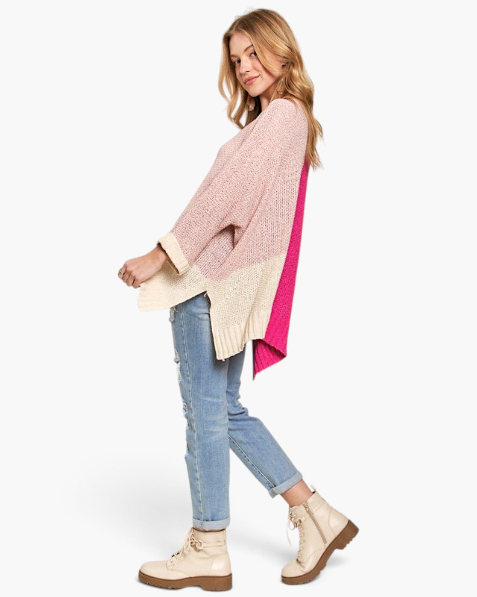 A woman modeling a color-block knit poncho sweater with light pink, cream, and bright pink panels, paired with light-wash blue jeans and beige lace-up ankle boots, against a plain white studio background.