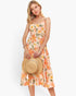 A woman modeling a sleeveless tiered midi dress with a peach, orange, and green floral print, holding a straw hat against a plain white studio background.