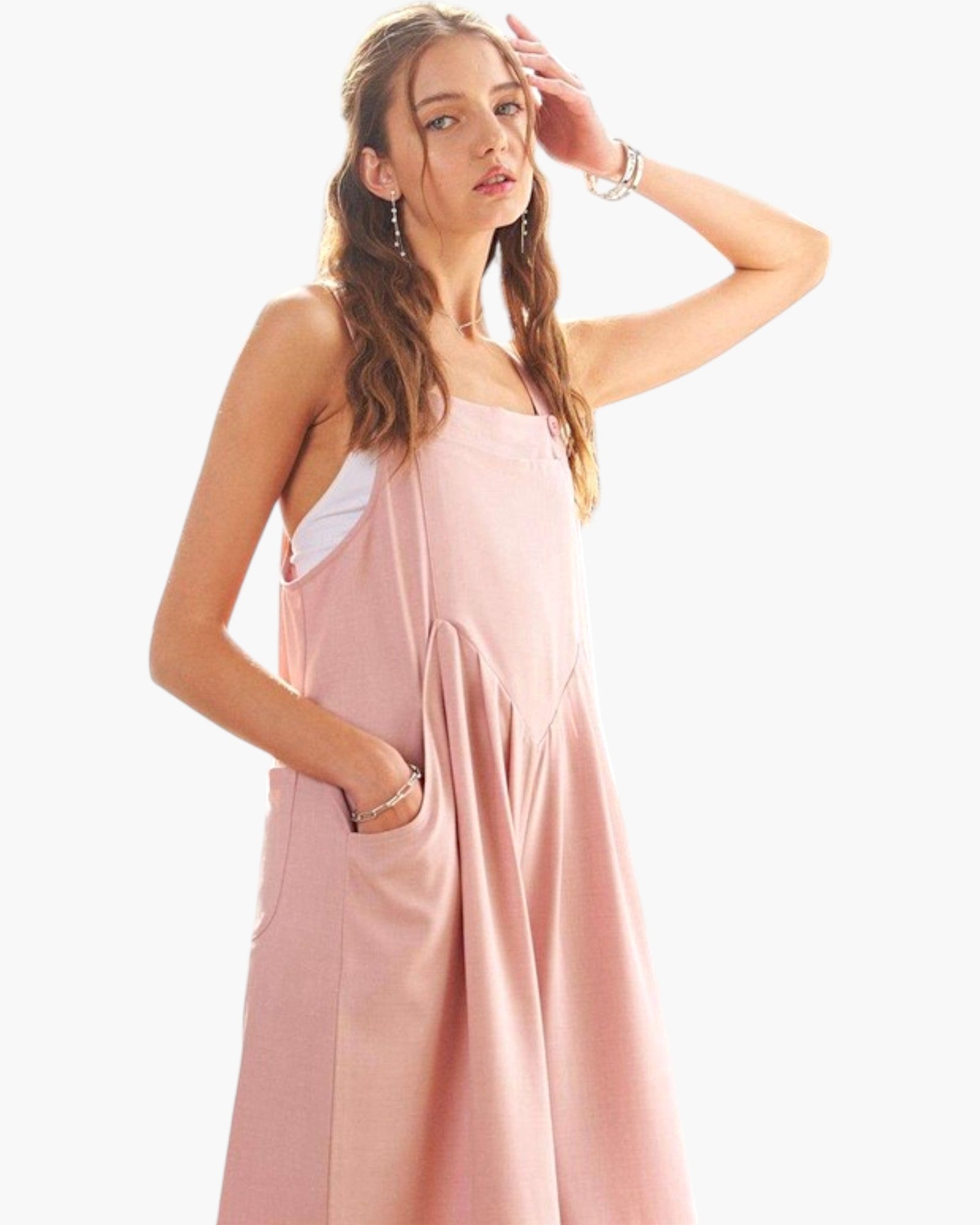 A woman modeling a light pink sleeveless pinafore-style dress with a white tank top underneath, posed against a clean white studio background.