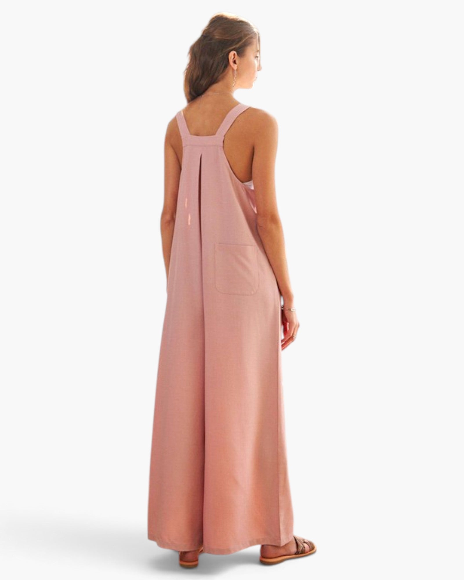 A woman viewed from the back wearing a dusty pink sleeveless jumpsuit with wide legs, a back pocket, and shoulder straps, paired with brown flat sandals against a plain white studio background.