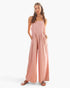 A woman modeling a dusty pink sleeveless jumpsuit with wide legs and side pockets, paired with brown sandals, against a plain white background.