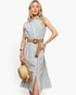 A woman models a sleeveless, button-front midi dress with vertical blue and white stripes, styled with a tan woven belt, holding a straw hat against a plain white background.
