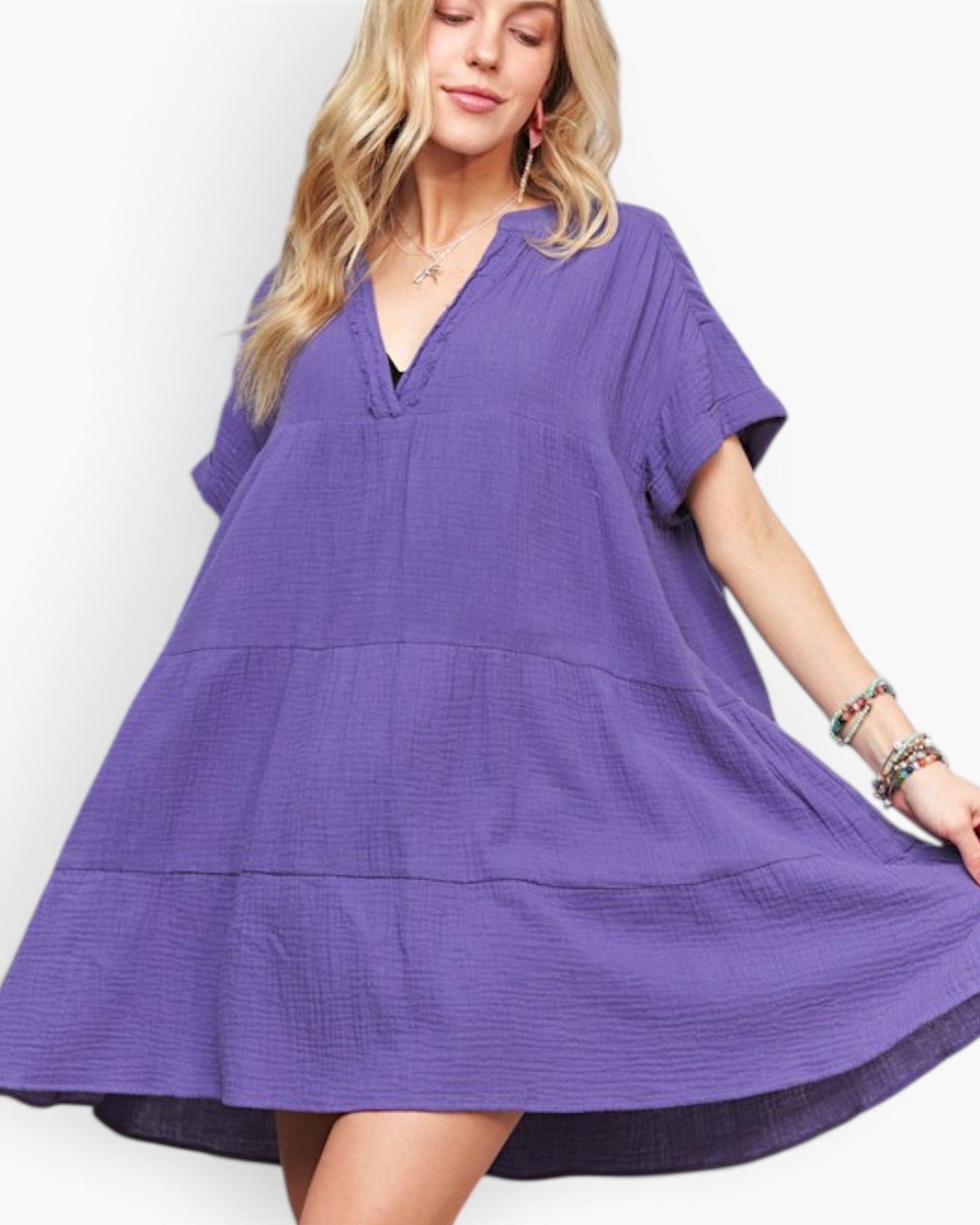 A woman wearing a loose, tiered purple dress with short sleeves and a split neckline, photographed against a clean white studio background.
