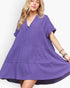 A woman wearing a loose, tiered purple dress with short sleeves and a split neckline, photographed against a clean white studio background.