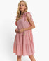 A woman modeling a pink tiered dress with a textured gingham pattern, featuring tie-shoulder straps and a small gold tassel charm at the waist, set against a plain white studio background.