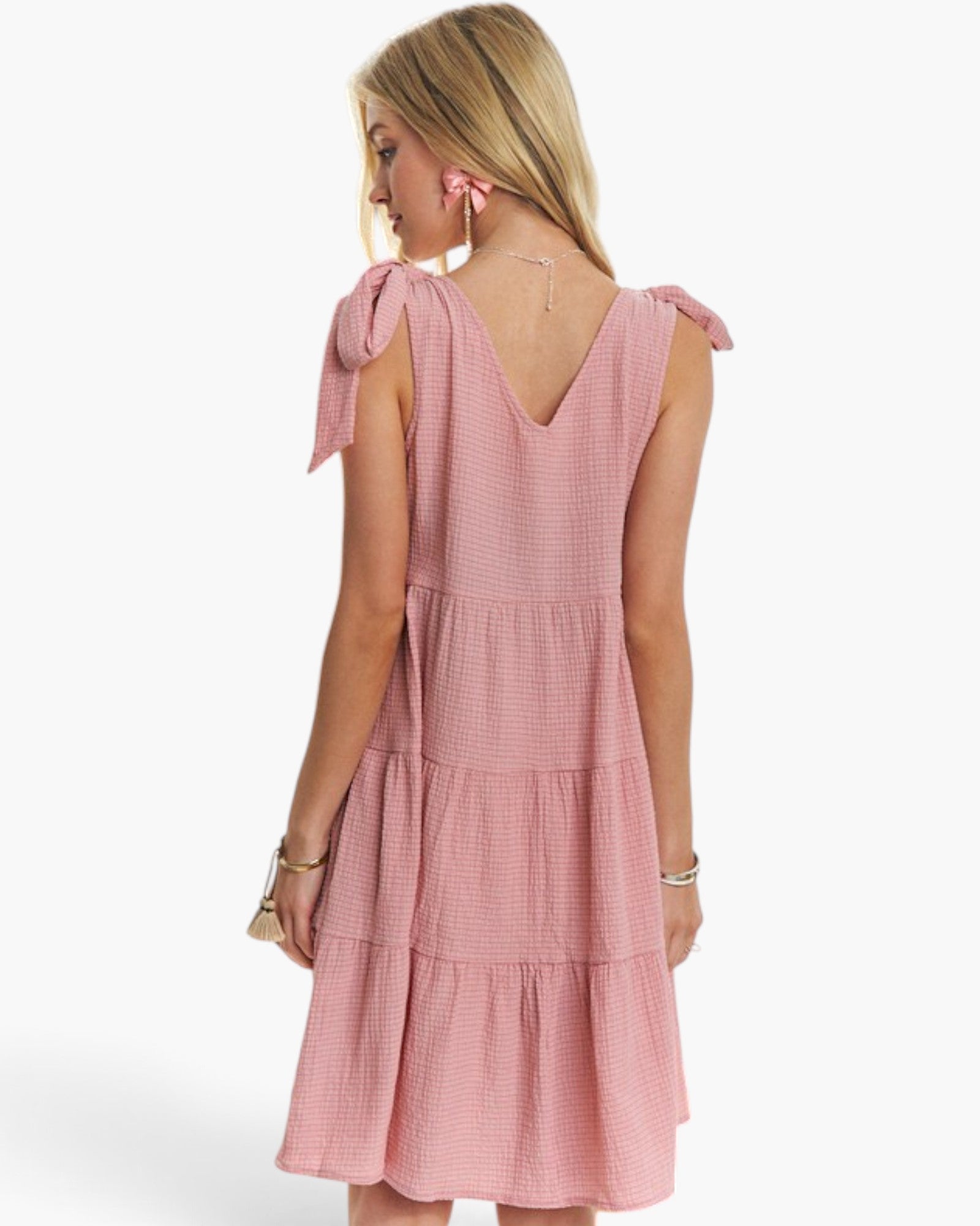 A woman shown from the back wearing a sleeveless pink tiered dress with bow details on the shoulders, photographed against a clean white studio background.