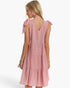 A woman shown from the back wearing a sleeveless pink tiered dress with bow details on the shoulders, photographed against a clean white studio background.
