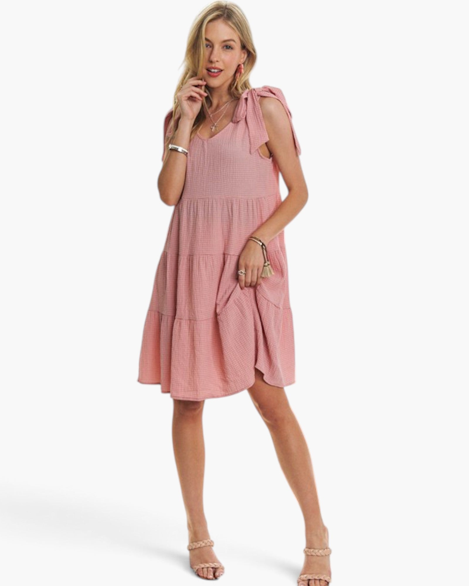 A woman modeling a pink tiered sleeveless dress with shoulder ties and side pockets, photographed against a clean white studio background.