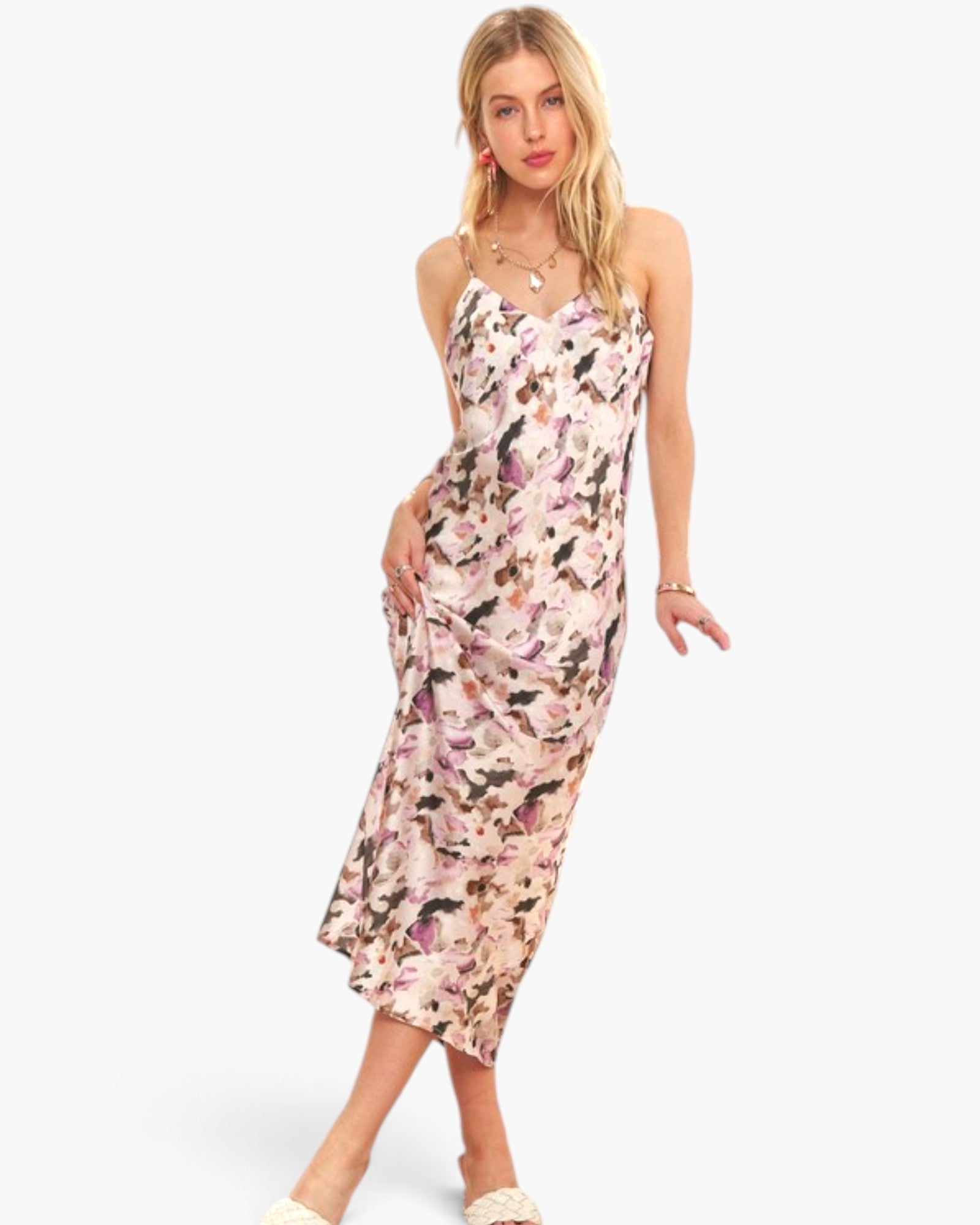 A woman modeling a sleeveless midi slip dress with a pink, purple, and black abstract watercolor pattern on a light beige background, set against a plain white studio backdrop.