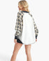 A woman shown from the back wearing a long-sleeve varsity-style jacket with a white back panel and black-and-beige patterned sleeves, paired with black shorts, against a light gray studio background.