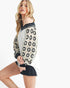 A woman modeling a white off-the-shoulder knit top with black and cream crochet patterned sleeves, paired with black denim shorts, against a plain white background.