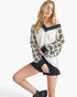 A woman modeling a long-sleeve off-the-shoulder top with a white body, black and beige paisley patterned sleeves, and black striped trim on the neckline and cuffs, paired with black denim shorts and black boots against a light gray studio background.
