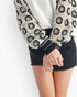 A close-up of a person wearing a cream crochet cardigan with black floral motifs, a white top, and a black denim mini skirt against a plain white background.