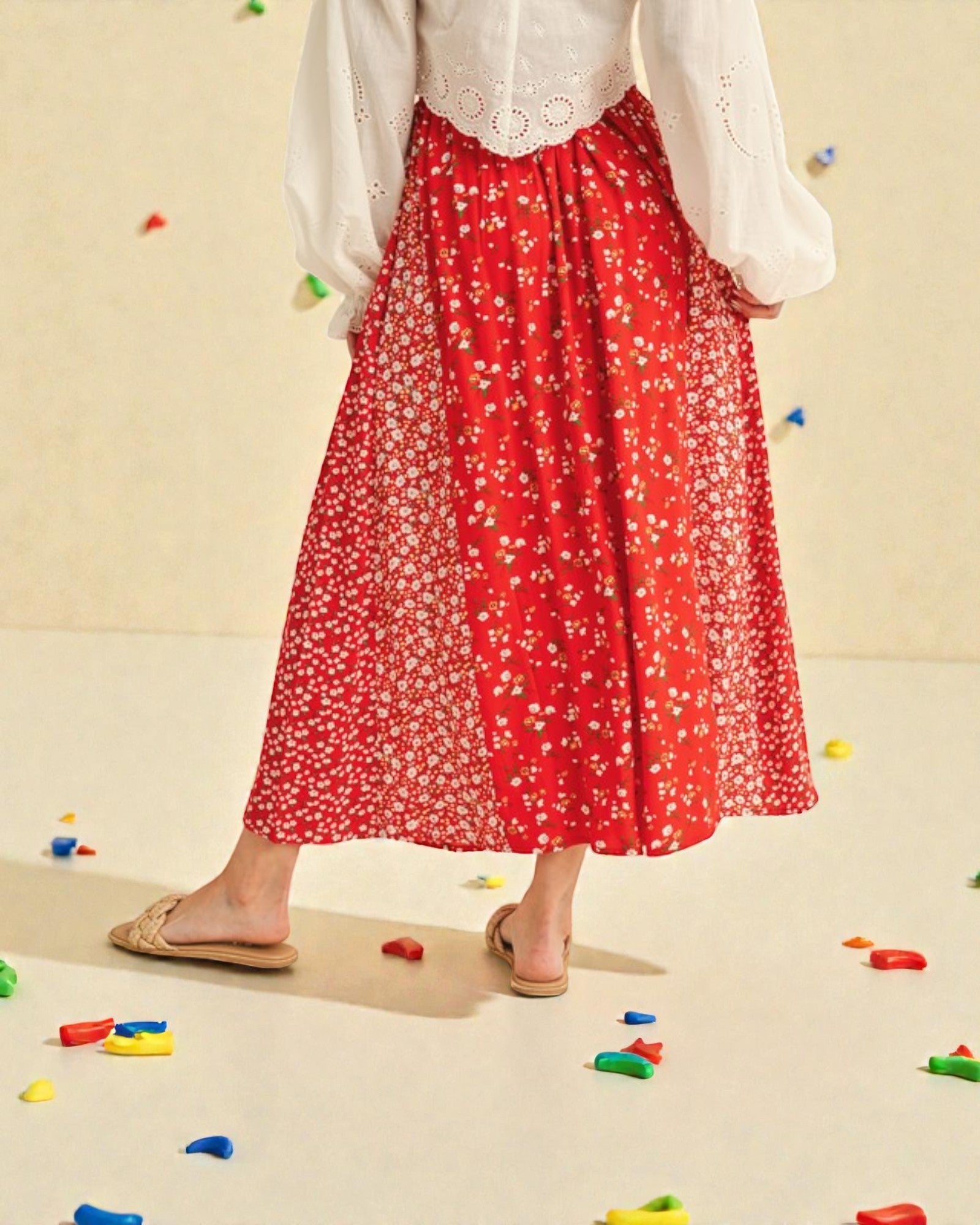 Person wearing a red floral skirt and white blouse on a beige background with colorful objects scattered around.