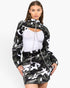 A model wearing a black and white camouflage zip-up jacket with a high collar and a matching mini skirt, styled over a white cutout top against a plain white background.