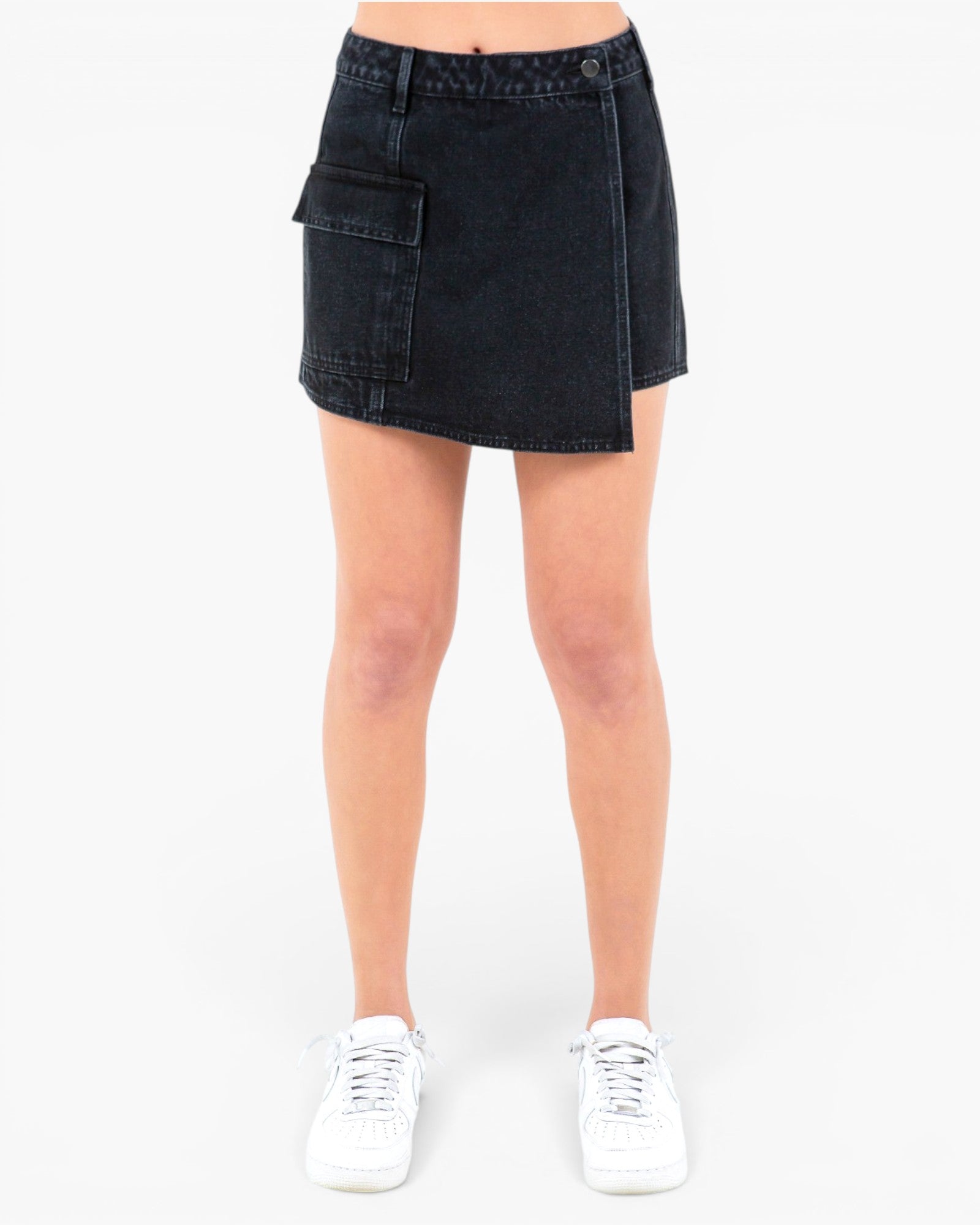 A model from the waist down wearing a dark wash denim mini skirt with a diagonal front slit and a large side cargo pocket, paired with white sneakers on a light gray background.