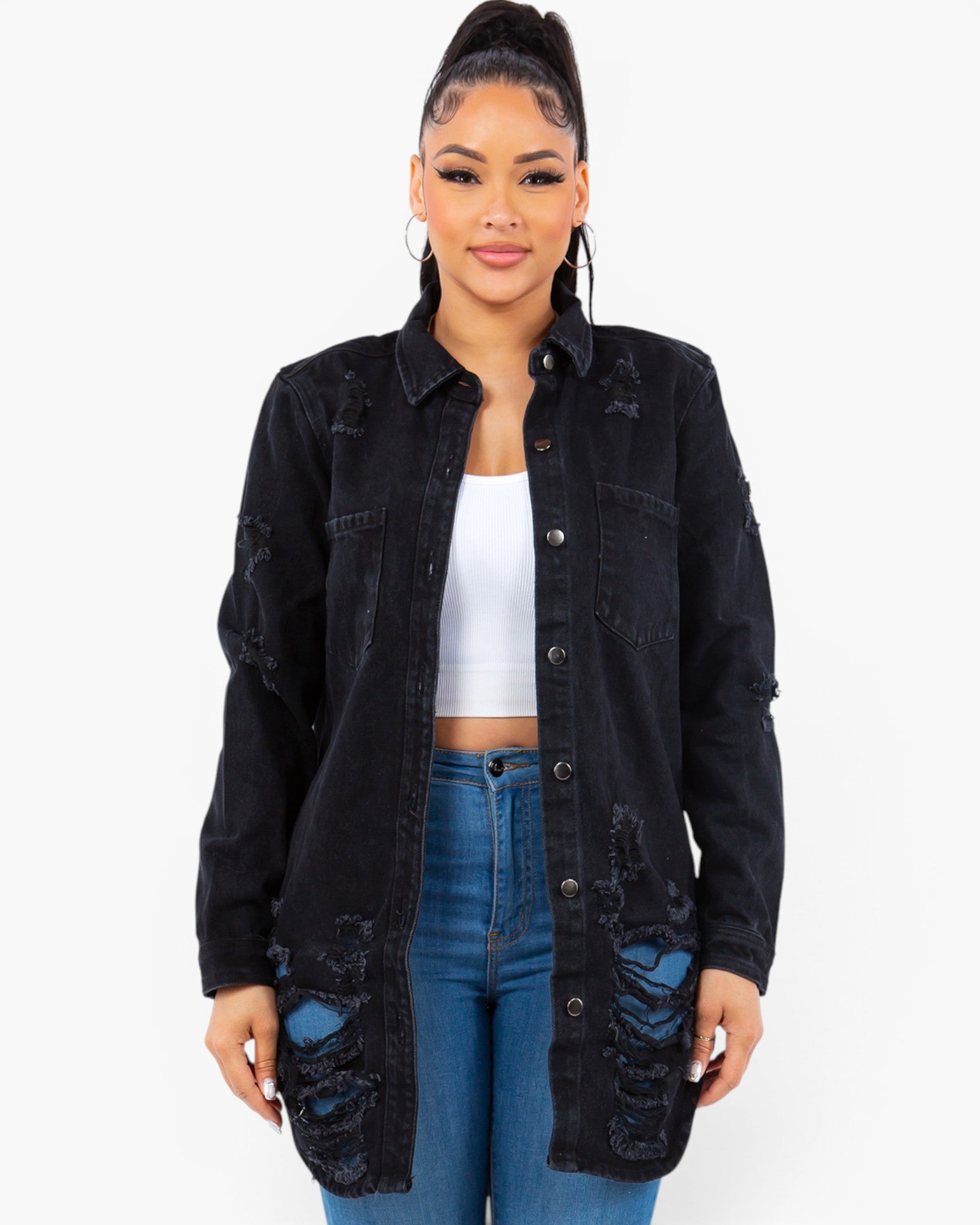 A woman modeling a black distressed denim jacket over a white crop top and blue jeans, photographed against a plain light gray studio background.