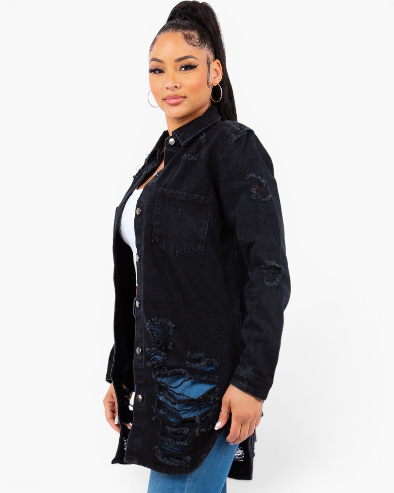 A woman modeling a black distressed denim shirt jacket with ripped details, worn over a white top and blue jeans, against a plain white background.