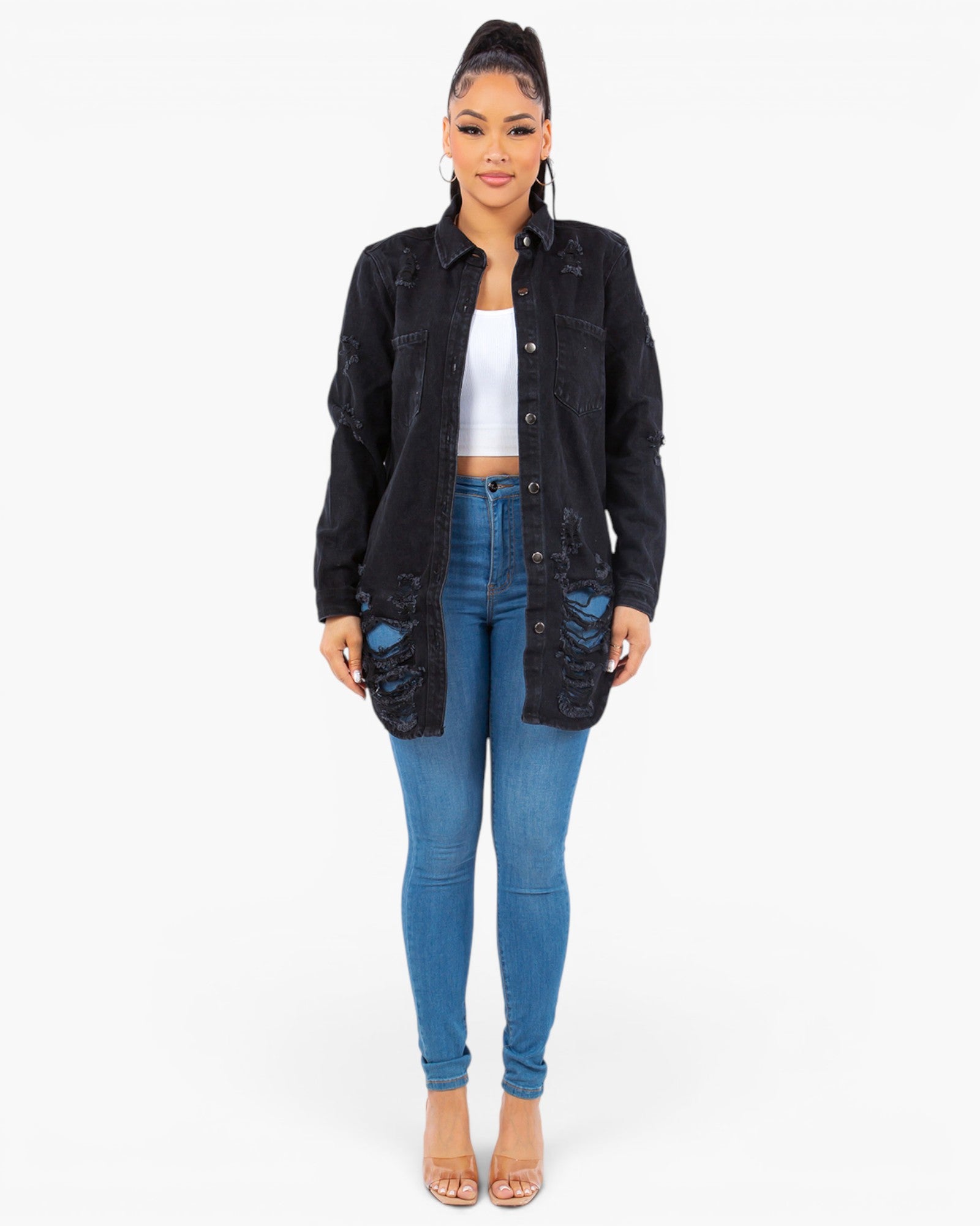 A woman modeling a black distressed denim jacket over a white crop top and blue skinny jeans, standing against a plain white studio background.