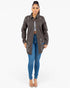 A woman modeling a dark brown distressed utility jacket with button details, worn over a white crop top and blue skinny jeans, against a plain white background.
