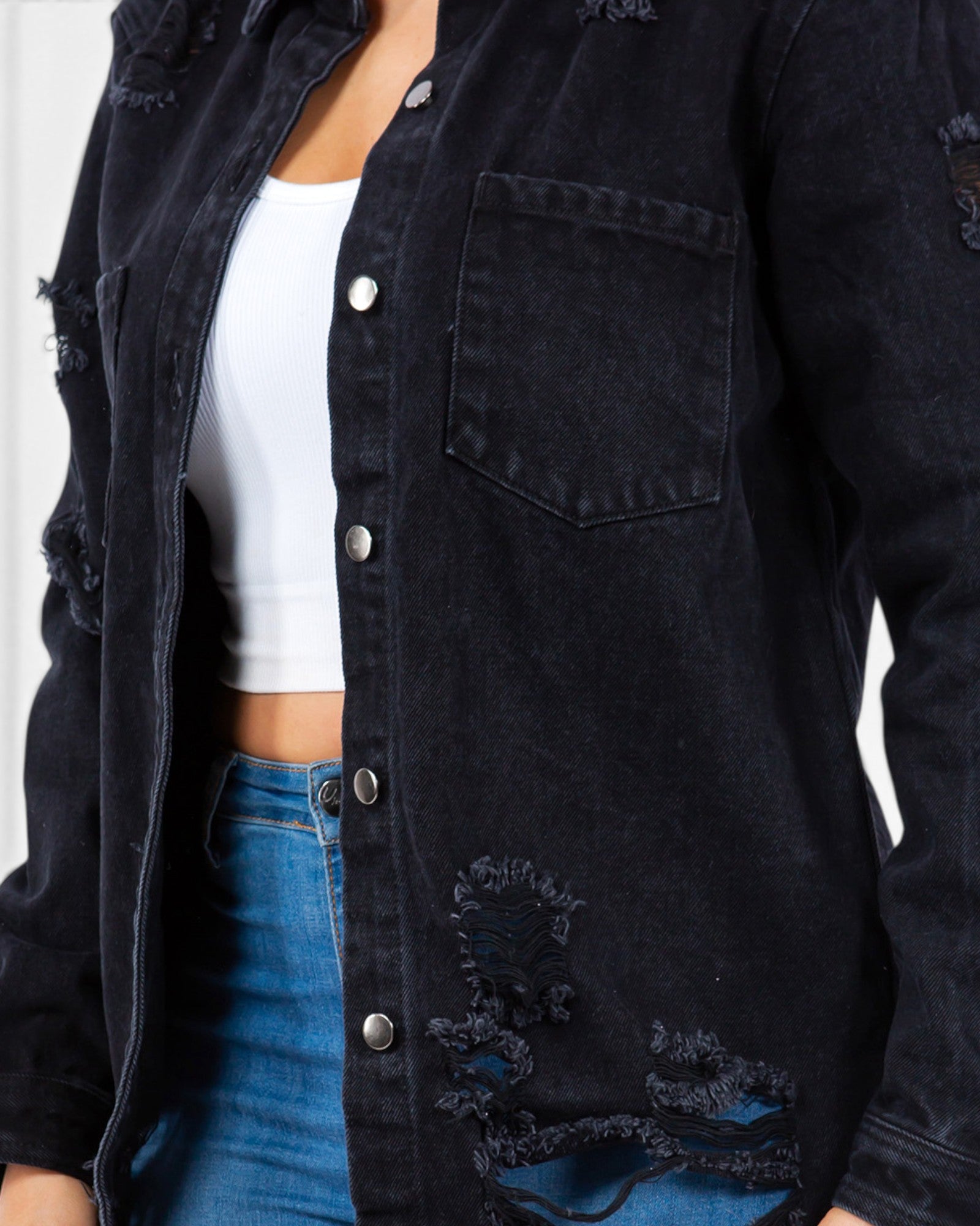 A close-up of a person wearing a dark washed distressed denim button-up shirt jacket over a white crop top and blue jeans.