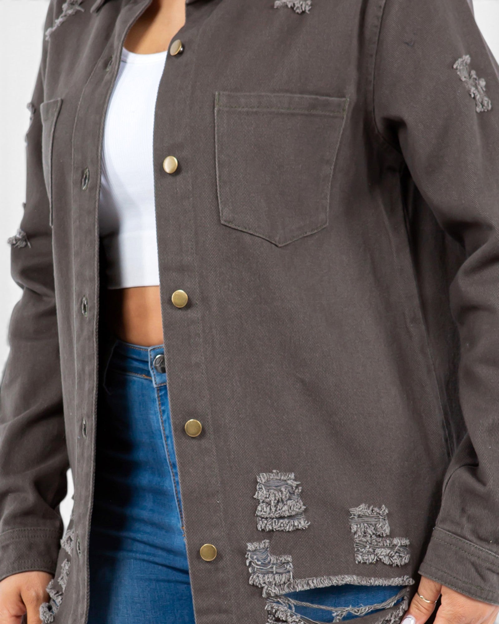 A close-up of a person wearing a dark gray distressed button-up shirt jacket over a white crop top and blue jeans, showing the chest pocket, brass-tone buttons, and frayed details.