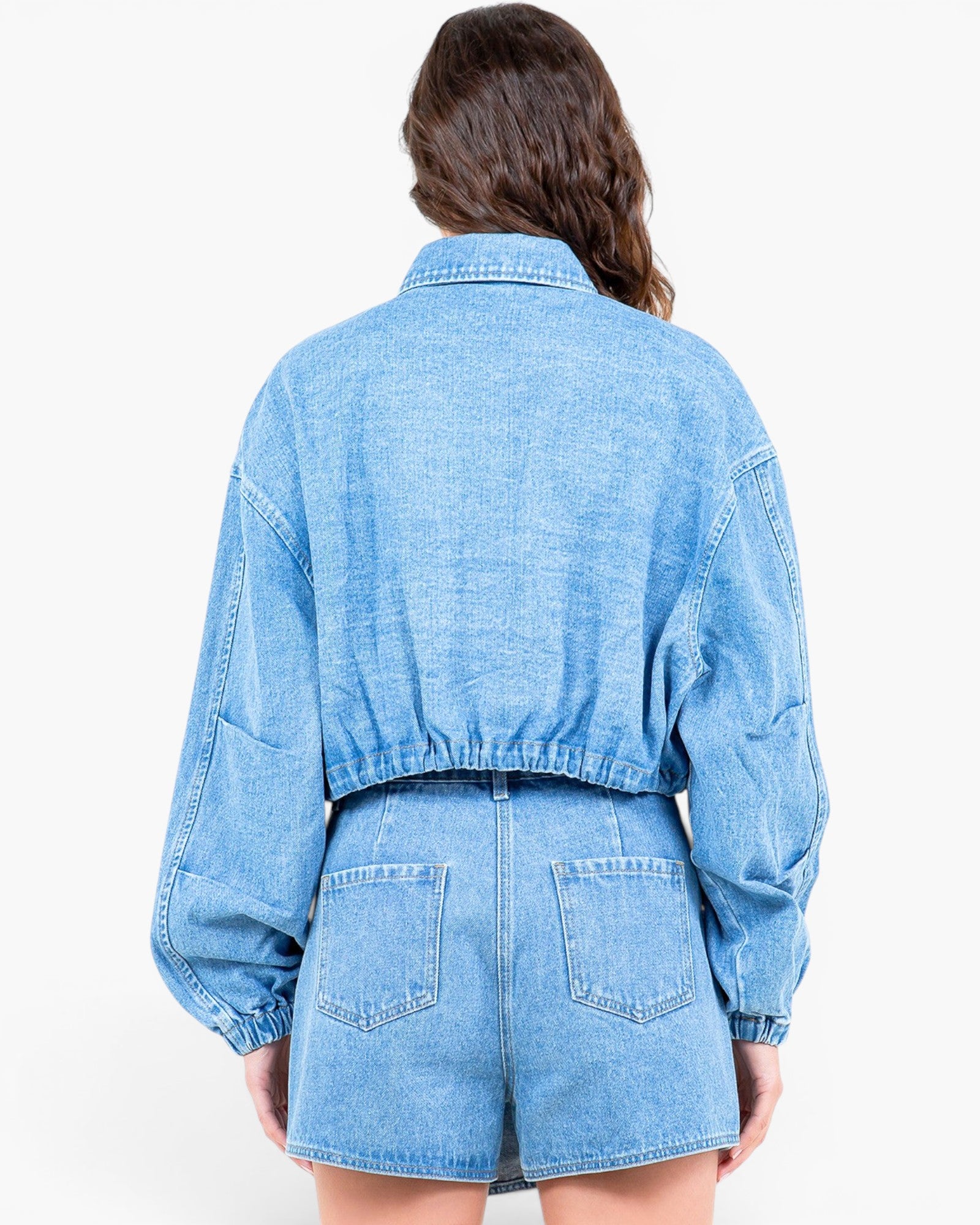 A model viewed from the back wearing a light blue denim jumpsuit with long balloon sleeves and matching shorts.