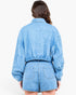 A model viewed from the back wearing a light blue denim jumpsuit with long balloon sleeves and matching shorts.
