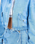 Close-up of a light blue denim jacket with a front zipper, flap chest pocket, and drawstring waist detail, worn over a white top.
