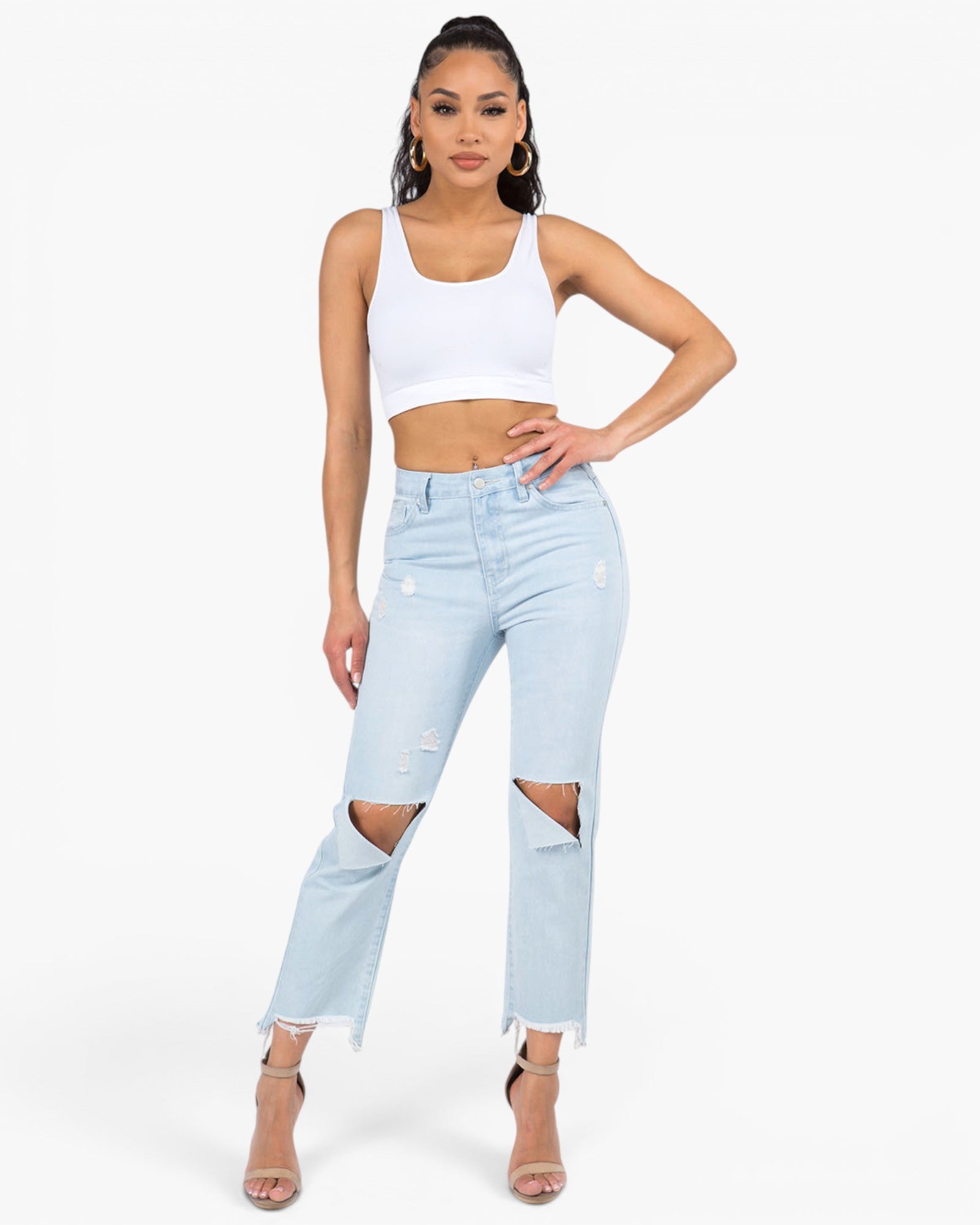 A woman modeling light-wash blue distressed jeans with large ripped knees, paired with a white crop top and nude high-heeled sandals, against a plain white studio background.