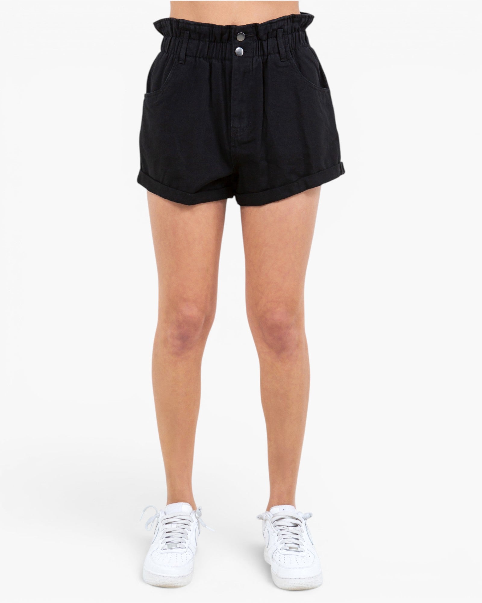 Black high-waisted shorts with a paperbag waistband and button closure, shown on a model from the waist down wearing white sneakers against a plain white background.