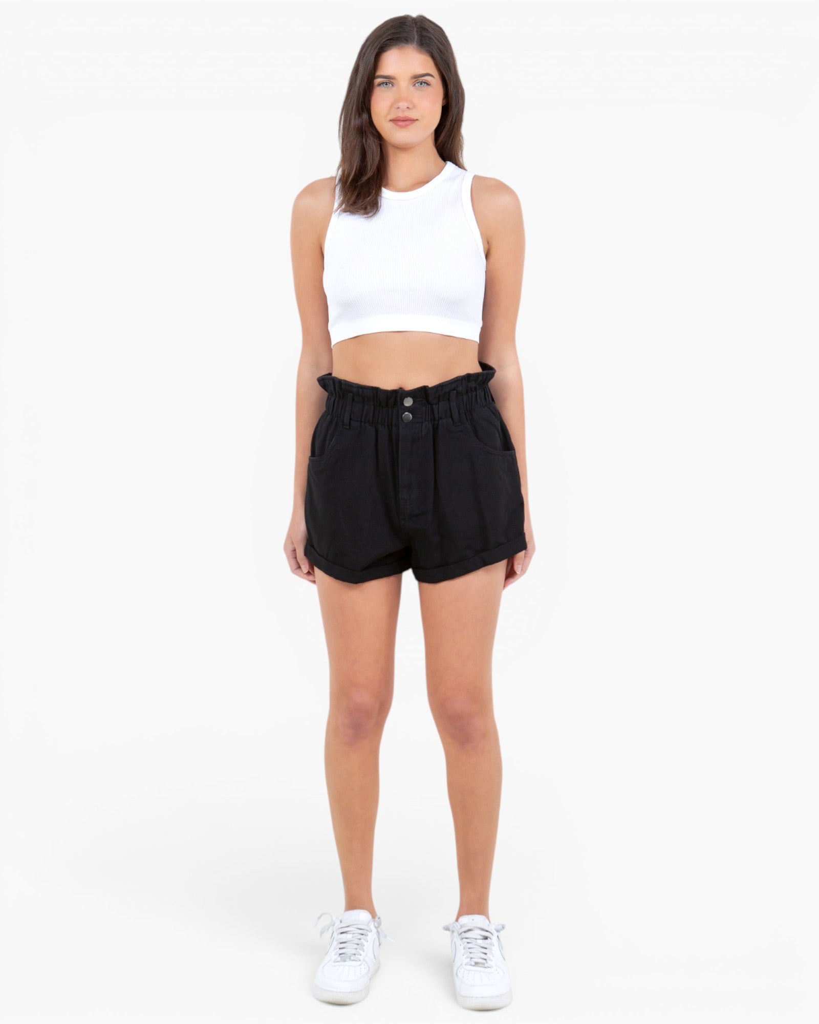 A woman modeling a white sleeveless crop top and black high-waisted shorts with white sneakers, standing against a plain white studio background.