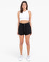 A woman modeling a white sleeveless crop top and black high-waisted shorts with white sneakers, standing against a plain white studio background.