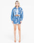A woman modeling a matching blue and white abstract patterned cropped jacket and mini skirt set, paired with a white crop top and nude high heels, against a plain white studio background.
