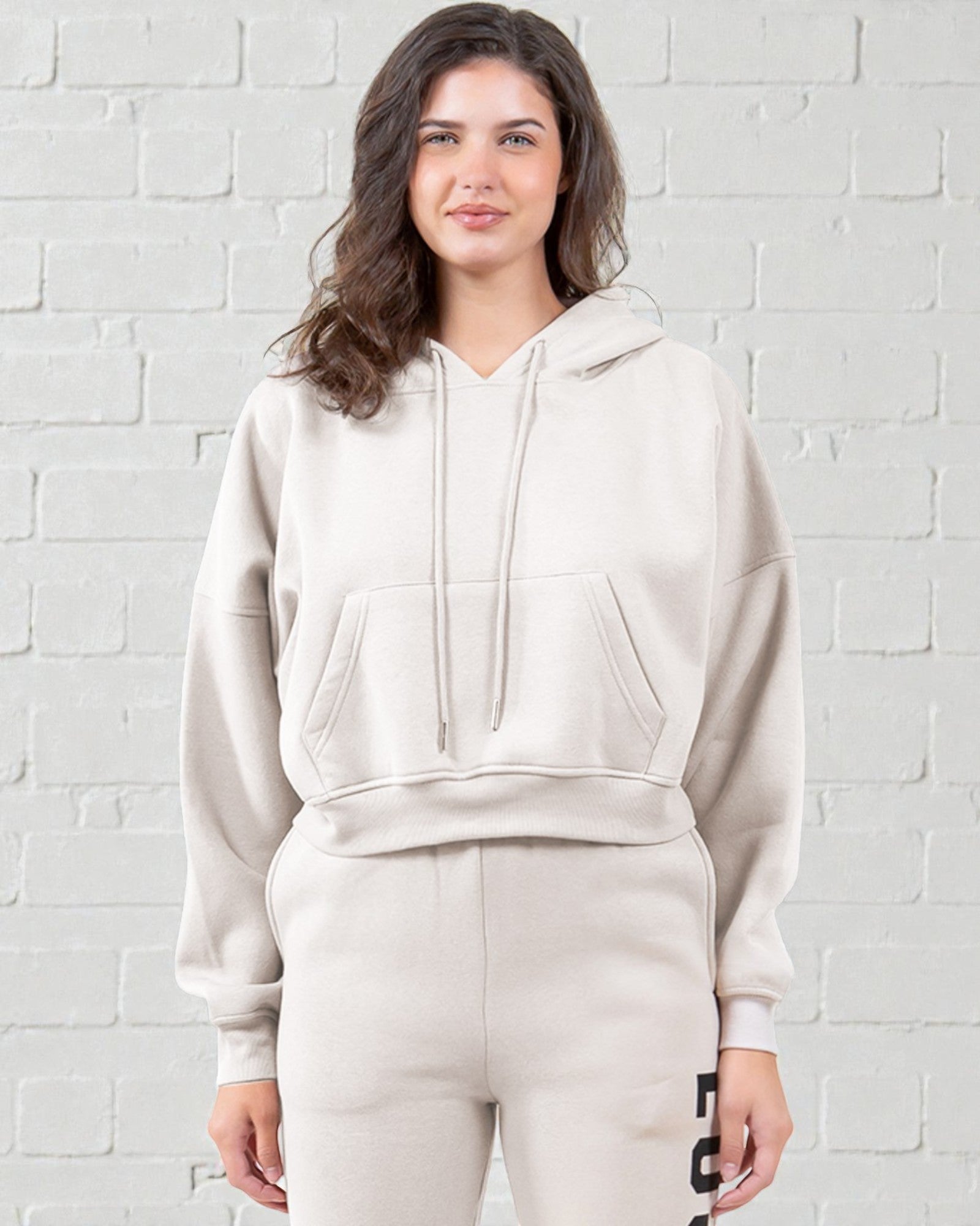 Woman wearing a beige hoodie and pants against a white brick wall