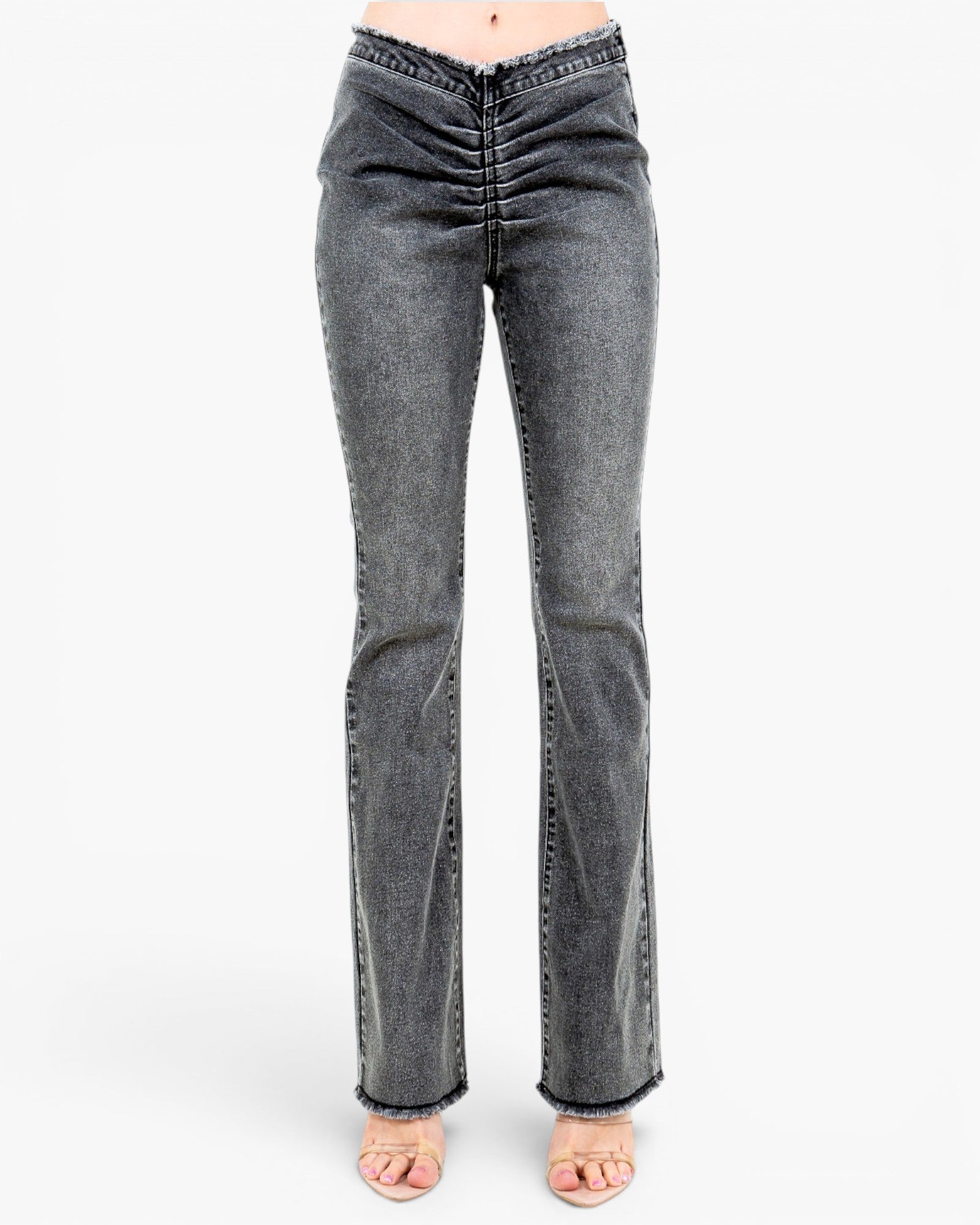 A pair of high-waisted, dark gray flared jeans with a ruched front and frayed hems, shown on a model wearing clear high-heel sandals against a white studio background.