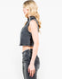 A woman shown in side profile wearing a cropped sleeveless black denim vest with raw frayed edges and matching dark gray jeans, against a plain white studio background.