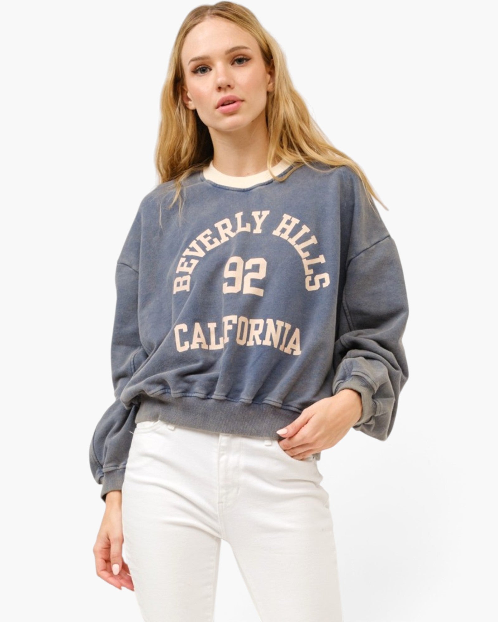 A woman modeling a faded blue-gray crewneck sweatshirt with 'BEVERLY HILLS 92 CALIFORNIA' printed in cream on the front, paired with white pants, against a plain white studio background.